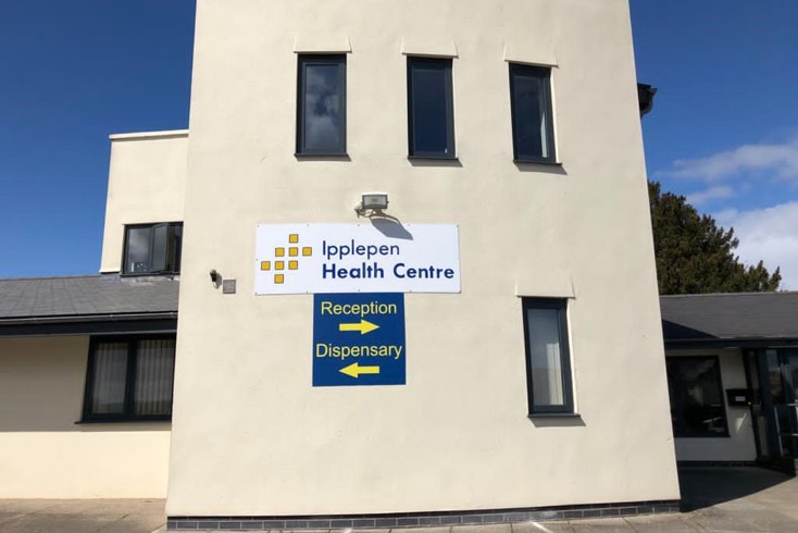 The exterior of Ipplepen health centre. There is a sign on the wall with an arrow pointing right to reception and an arrow pointing left to dispensary. 